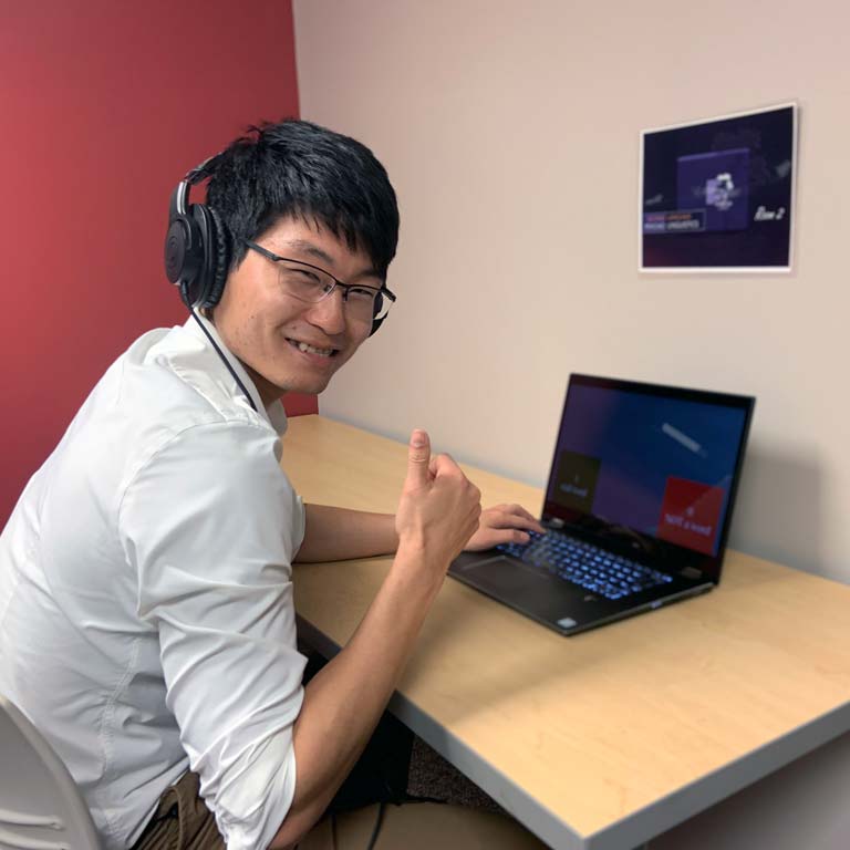 Student at computer wearing glasses and headphones, smiling and holding up a thumbs-up